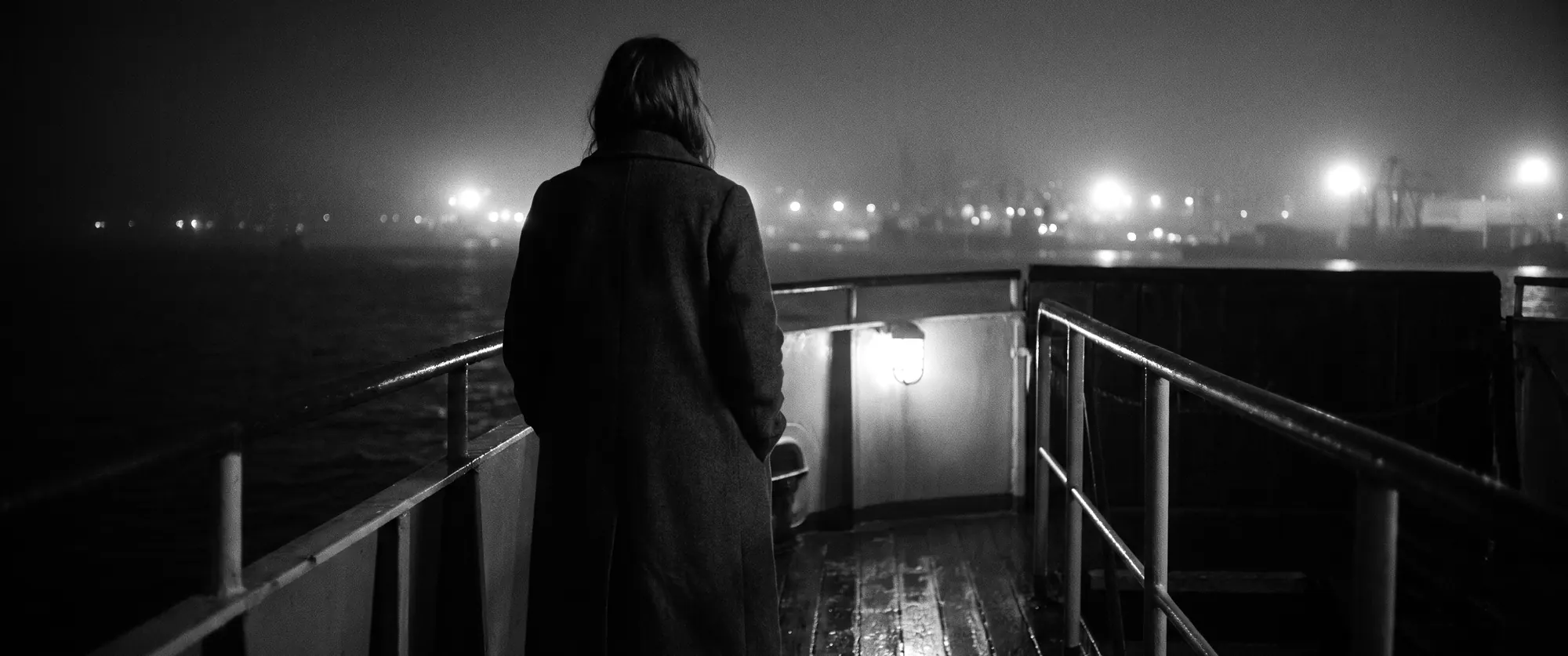 A woman in a long coat at the rail of a night ferry, harbour lights in the fog