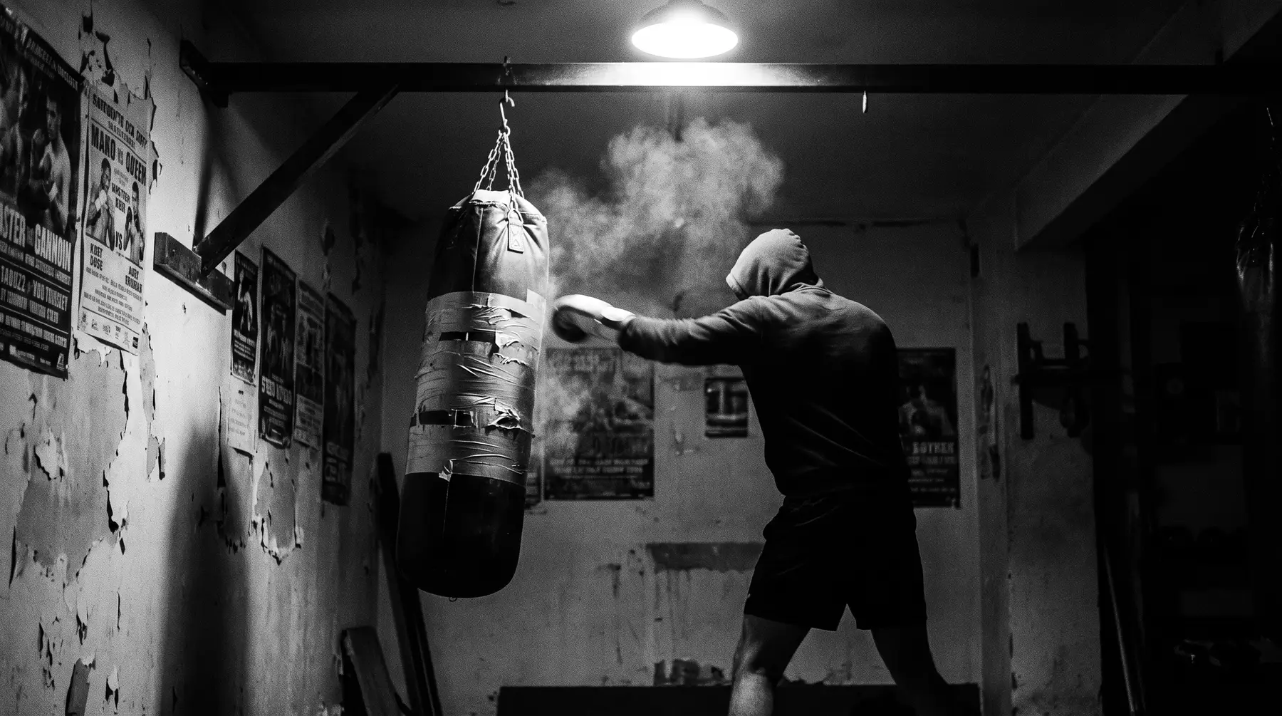 A boxer in a hood drives a punch into a heavy bag under one bare bulb, chalk dust in the air
