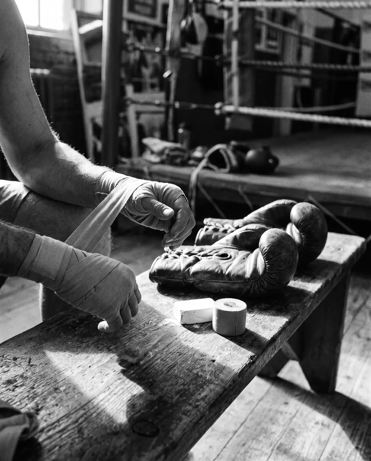 Hands being wrapped on a wooden bench, worn gloves and tape beside them